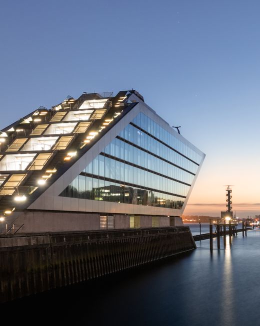 A modern multi-storey commercial building with a prominent curved glass roof section at the top, featuring a red accent line along the edge. The building has a facade of grey panels and large reflecti
