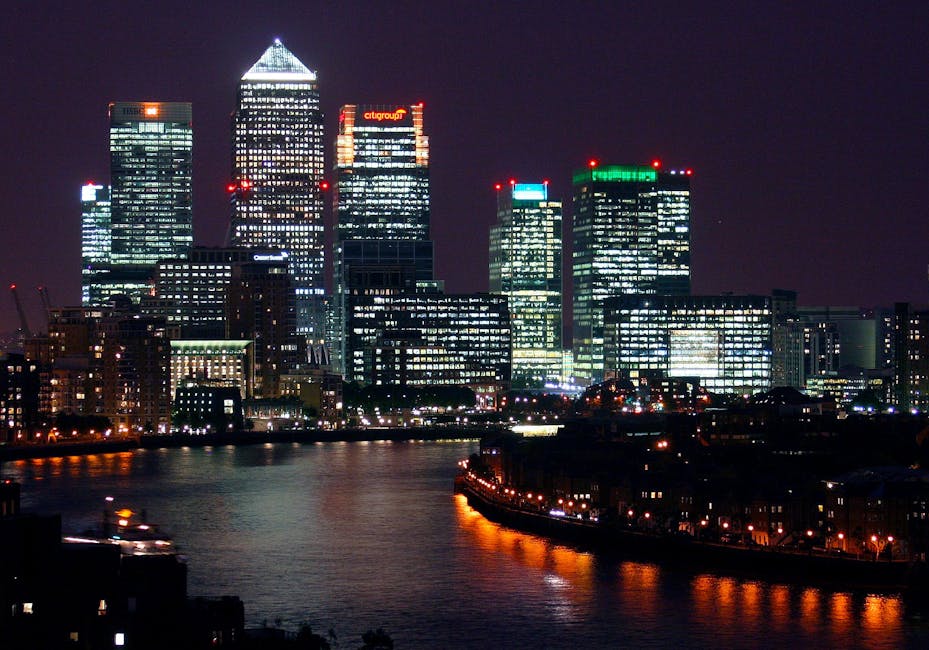 A nighttime cityscape of Canary Wharf in London's Docklands, featuring illuminated skyscrapers with modern glass facades reflecting city lights on the River Thames in the foreground. Multiple high-rise office buildings display bright internal lighting and exterior red and green aviation warning lights. The scene captures the vibrant urban environment associated with house removals and home relocation logistics, with the skyline providing a relevant backdrop for commercial and residential moving services such as those offered by Man and Van Docklands.