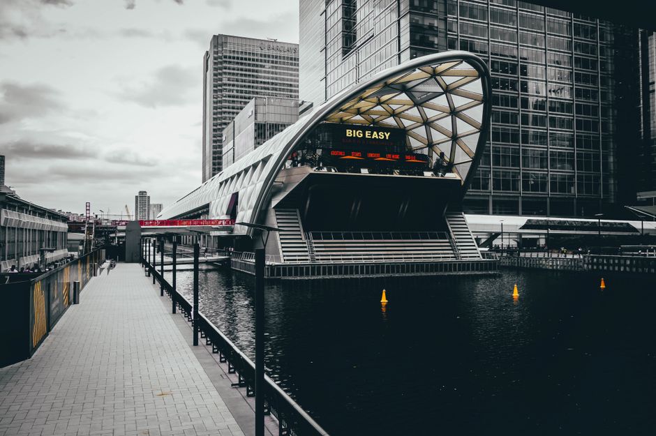A modern, curved architectural structure extends over a waterway in Canary Wharf, with a large digital display screen showing the message 'BIG EASY' alongside a scrolling ticker update. The foreground features a paved walkway with a black metal railing, leading towards the building, which is surrounded by tall glass skyscrapers reflecting the overcast sky. Inside or near the building, there are visible cardboard boxes wrapped in clear plastic and packed with household items, some stacked on a trolley for transport. A man and van service, such as Man and Van Docklands, might be involved in home relocation or furniture transport in this area, focusing on efficient packing and loading processes. The image captures the environment during daylight, emphasizing the urban setting typical of city-based removals and moving logistics, with a focus on the environment conducive to tight access moves often associated with relocating in the Docklands district.