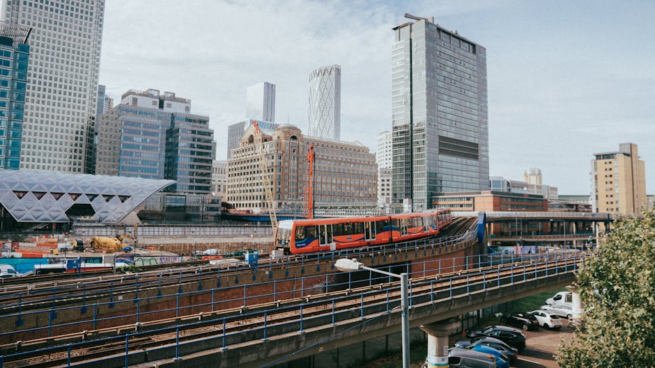 Photograph of a cityscape featuring modern high-rise office buildings with glass exteriors reflecting the sky, situated along a river in Canary Wharf. A red and white train is visible moving along the