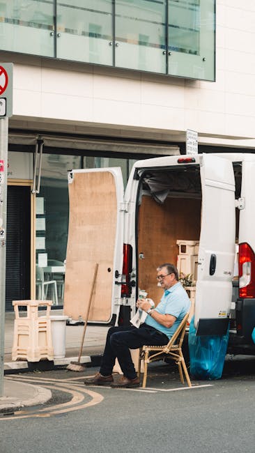A man wearing glasses and a light blue shirt sits on a wooden chair beside an open white van used for house removals, parked on a city street. The van's rear doors are wide open, revealing an interior filled with various packing materials and boxes prepared for a home relocation. A large wooden panel leans against the van's side, with a small pallet nearby on the pavement. The scene takes place in front of a modern building with glass windows and a beige facade. The street includes a no-parking sign and markings on the road indicating a designated loading area, suggesting the man and van, part of a professional removals service like Man and Van Docklands, are engaged in furniture transport and packing and moving logistics, focusing on efficient loading procedures in narrow street environments typical of the Docklands area.