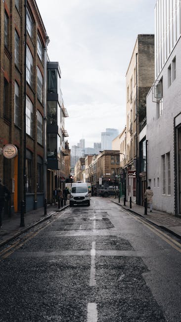 A narrow street scene in the Isle of Dogs area, showing modern and older multi-storey buildings lining both sides of the paved road. The street appears wet, possibly from recent rain, and is marked with a central dashed line. On the left side, there are storefronts with large windows, and on the right, a white building with several small windows. A white van is parked partially on the pavement near the center of the image, indicating a moving or loading activity. In the background, taller office or residential buildings are visible, with a cloudy sky overhead. The scene is well-lit with natural daylight, illustrating an urban environment suitable for local removals and home relocation logistics, as handled by companies like Man and Van Docklands, particularly relevant for navigating narrow streets during furniture transport or packing and moving operations.