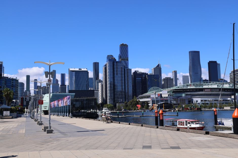 A view of a modern city skyline with tall glass skyscrapers against a clear blue sky, situated across a body of water with leisure boats docked along the quay. In the foreground, there is a paved prom